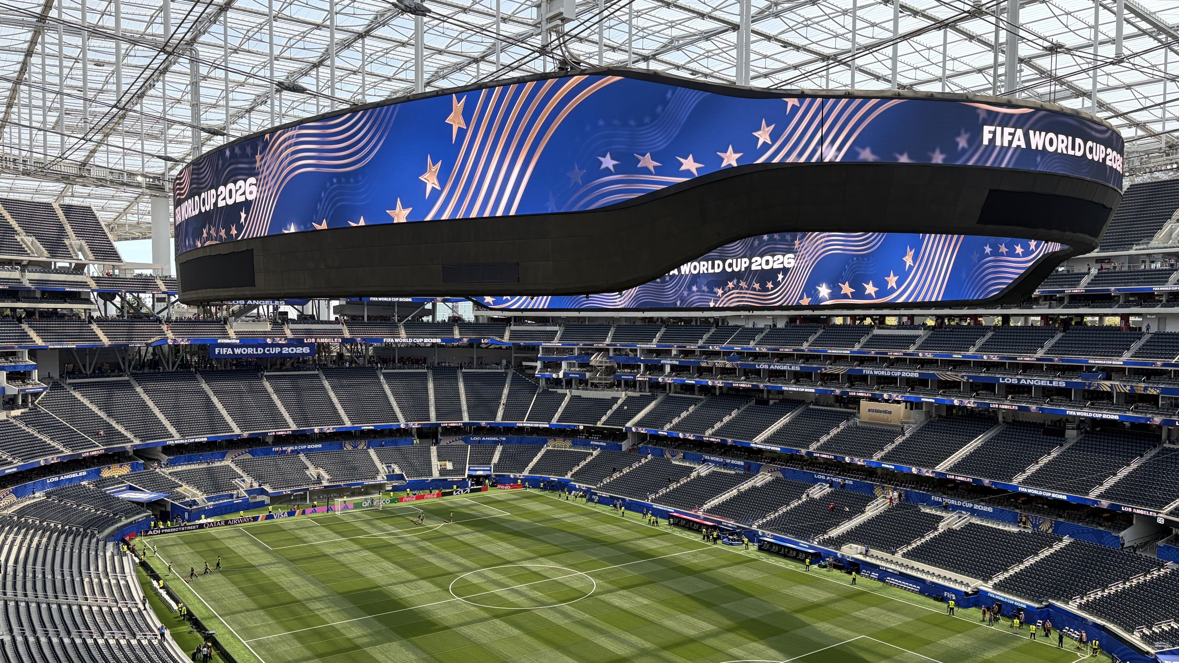 The interior of SoFi Stadium three hours before a 2026 FIFA World Cup match, the central scoreboard reading FIFA World Cup 2026 above an empty pitch.